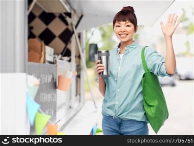 sustainability, eco living and people concept - asian woman with thermo cup for hot drinks and green reusable shopping bag waving hand over food truck background. woman with tumbler and bag over food truck