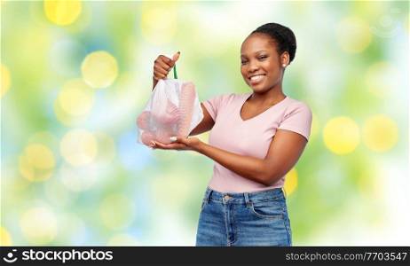 sustainability, eco living and people concept - portrait of happy smiling african american woman holding reusable net bag with vegetables over lights on green background. happy woman with vegetables in reusable net bag