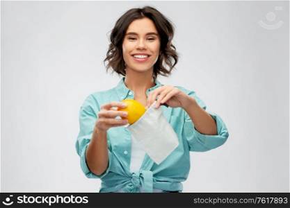 sustainability, eco living and people concept - portrait of happy smiling young woman in turquoise shirt putting orange into reusable bag over grey background. happy woman putting orange into reusable bag