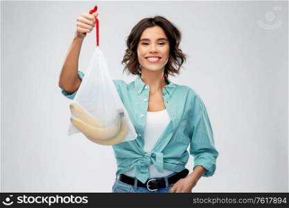 sustainability, eco living and people concept - portrait of happy smiling young woman in turquoise shirt and jeans holding reusable string bag with bananas over grey background. happy woman with bananas in reusable string bag