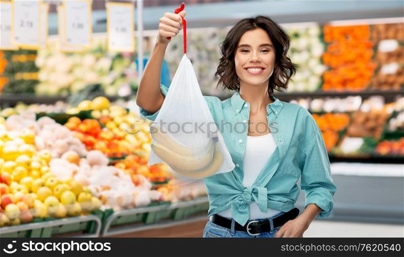 sustainability, eco living and people concept - portrait of happy smiling young woman in turquoise shirt and jeans holding reusable string bag with bananas over grocery store or supermarket background. happy woman with bananas in reusable string bag