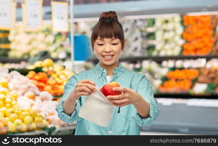 sustainability, eco living and people concept - portrait of happy smiling young asian woman in turquoise shirt putting apple into reusable bag over grocery store background. happy woman putting apple into reusable bag