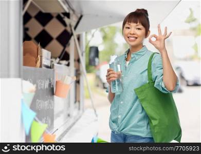 sustainability, eco living and people concept - portrait of happy smiling young asian woman with green reusable shopping bag and glass bottle of water showing ok gesture over food truck background. woman with shopping bag and bottle over food truck