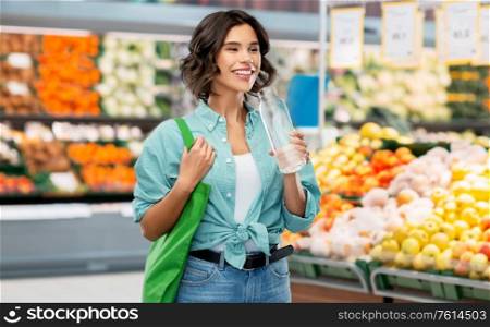sustainability, eco living and people concept - portrait of happy young woman with green reusable canvas bag for food shopping and glass bottle of water over supermarket or grocery store background. woman with bag for food shopping and glass bottle