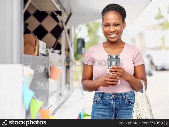 sustainability, eco living and people concept - smiling african american woman with thermo cup or tumbler for hot drinks and string bag over food truck background. woman with tumbler and string bag ober food truck