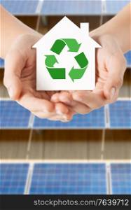 sustainability, energy saving and consumption concept - close up of hands holding house with green recycling symbol over solar panels on background. hands holding house with recycling symbol