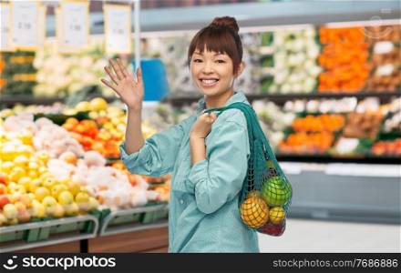 sustainability, food shopping and eco friendly concept - happy smiling asian woman in turquoise shirt holding reusable string bag with fruits and vegetables waving hand over grocery store background. asian woman with food in reusable bag at store
