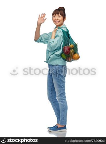 sustainability, food shopping and eco friendly concept - happy smiling asian woman in turquoise shirt and jeans holding reusable string bag with fruits and vegetables waving hand over white background. happy asian woman with food in reusable string bag