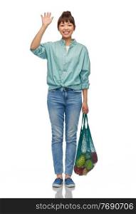 sustainability, food shopping and eco friendly concept - happy smiling asian woman in turquoise shirt and jeans holding reusable string bag with fruits and vegetables waving hand over white background. happy asian woman with food in reusable string bag