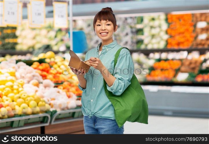 sustainability, food shopping and eco friendly concept - happy smiling asian woman in turquoise shirt and jeans holding reusable green tote bag and takeaway wok over grocery store background. asian woman with reusable bag for food and wok