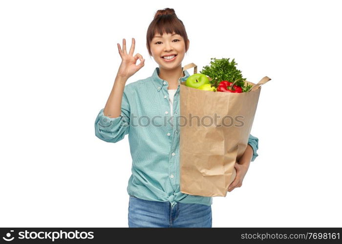 sustainability, food shopping and eco friendly concept - happy smiling asian woman in turquoise shirt and jeans holding paper bag with fruits and vegetables showing ok gesture over white background. happy asian woman with food in paper shopping bag