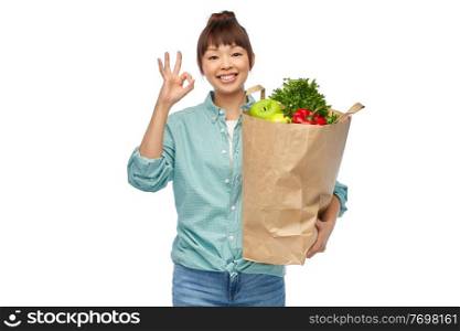 sustainability, food shopping and eco friendly concept - happy smiling asian woman in turquoise shirt and jeans holding paper bag with fruits and vegetables showing ok gesture over white background. happy asian woman with food in paper shopping bag