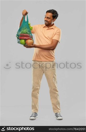 sustainability, food shopping and eco friendly concept - happy smiling indian man holding reusable net tote with fruits and vegetables on grey background. happy indian man with food in reusable net tote