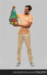 sustainability, food shopping and eco friendly concept - happy smiling indian man holding reusable net tote with fruits and vegetables on grey background. happy indian man with food in reusable net tote