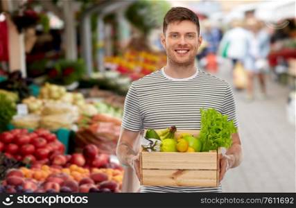sustainability, food shopping and eco friendly concept - happy smiling man with fruits and vegetables in wooden box over street market on background. happy man with food in wooden box at street market