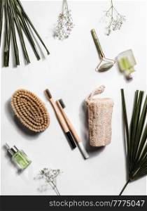 Sustainable bath and skin care products on white background with palm leaves: jade massage roller, bamboo toothbrush, massage brush, glass cosmetic bottle and natural sponge. Top view