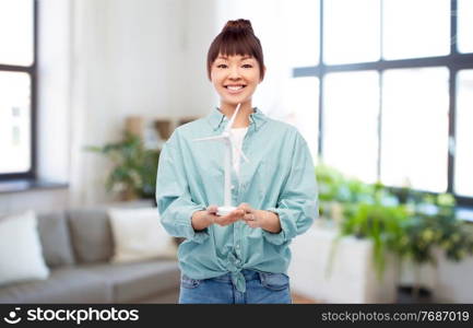 sustainable energy, power and people concept - happy smiling young asian woman with toy wind turbine over home background. smiling young asian woman with toy wind turbine