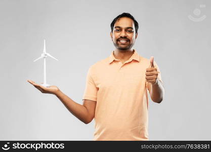 sustainable energy, power and people concept - happy smiling young indian man with toy wind turbine showing thumbs up over grey background. indian man with toy wind turbine showing thumbs up