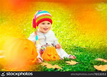 Sweet baby boy wearing cute colorful hat sitting on fresh green grass, little child sitting near two beautiful carved pumpkins, happy Halloween holiday