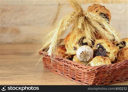 Sweet buns with poppy seeds lie in a wicker basket, on a wooden table and near a stone wall - sandstone. Poppy head and spikelets lie on sweet rolls. Sweet rolls with poppy seeds lie in a wicker basket