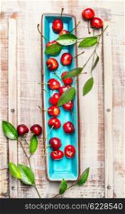 Sweet cherries with green leaves on blue rectangular plate on light wooden rustic background, top view. Summer fruits and berries concept.