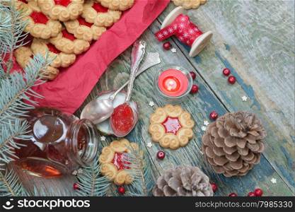 Sweet Christmas cookies with jam, fir branches and cones, cranberries, red burning candle and the bobbin with decorative ribbon on the old wooden table