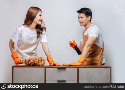 Sweet couple helping to cleaning furniture at home, Young man and woman wearing apron and rubber gloves use spray bottle and feather duster with happiness together