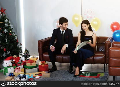 Sweet couple Love smile and spending Romantic christmas time and celebrating new year eve on Brown Sofa decoration with Christmas tree, colorful balloon and Gift Boxes in Living room at home