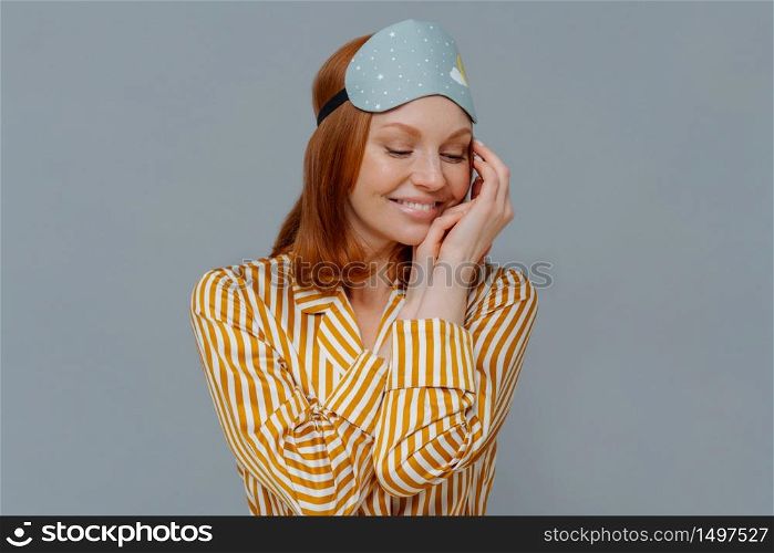 Sweet dreams and good rest concept. Pretty redhead woman turns head aside, keeps gaze down and touches cheek gently, wears striped pajama, blindfold, smiles positively, isolated over grey wall
