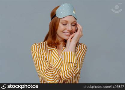 Sweet dreams and good rest concept. Pretty redhead woman turns head aside, keeps gaze down and touches cheek gently, wears striped pajama, blindfold, smiles positively, isolated over grey wall