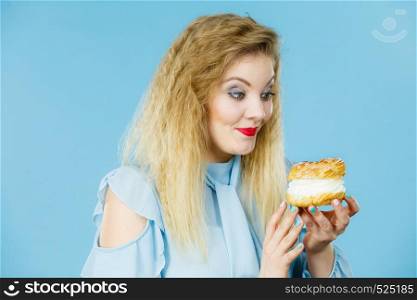 Sweet food and happiness concept. Funny joyful blonde woman holding yummy choux puff cake with whipped cream, excited face expression. On blue. Funny woman holds cream puff cake