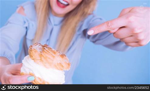 Sweet food and happiness concept. Funny joyful blonde woman pointing on yummy choux puff cake with whipped cream, excited face expression. On blue. Funny woman holds cream puff cake