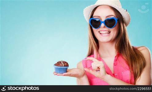 sweet food happiness and people concept. Smiling summer fashionable woman wearing straw hat heart shaped sunglasses holds cake cupcake in hand blue background
