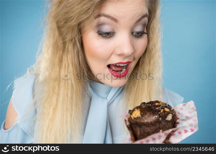 Sweet food sugar make us happy. Attractive blonde woman holds yummy chocolate cupcake in hand, open mouth, craving for cake. On blue. Attractive woman holds cake in hand