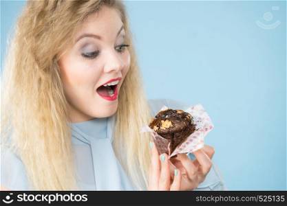 Sweet food sugar make us happy. Attractive blonde woman holds yummy chocolate cupcake in hand, open mouth, craving for cake. On blue. Attractive woman holds cake in hand