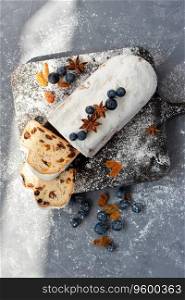 Sweet fruit cake decorated with raisins, blueberry and sugar powder icing, traditional Christstollen, stollen sliced on pieces, on a gray table with sunlight shadows. Sweet fruit cake decorated with raisins, blueberry and sugar powder icing, traditional Christstollen, stollen sliced on pieces, on gray table with sunlight shadows