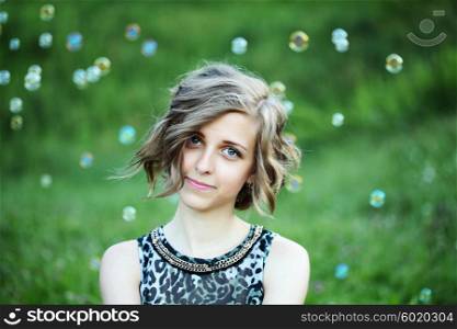 Sweet, happy, smiling blonde girl sitting on a grass in a park playing with bubbles and laughing, having fun. Lifestyle, outdoors