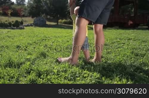 Sweet infant baby boy taking first steps barefoot with the help of affectionate father on green park lawn. Back view. Low view. Cute toddler child with caring dad learning to walk on grassy field in summer park. Slow motion. Steadicam stabilized shot