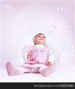 Sweet little angel sitting at the studio and playing with soap bubbles, having fun, wearing white fluffy wings, happy childhood concept