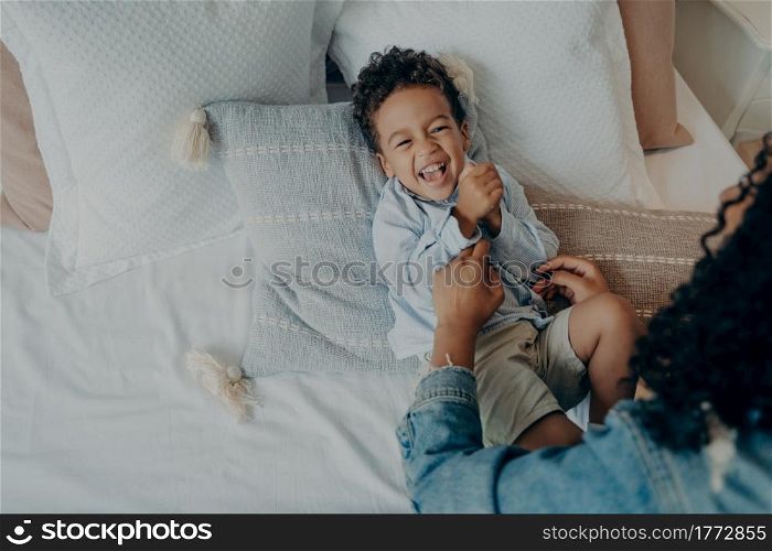 Sweet little boy kid laughing loudly and smiling while his loving mom tickling him on bed with lot of soft pillows, mother telling really funny story to son while spending happy time together at home. Sweet excited mixed race boy lying on bed and laughing while playing with his loving mom