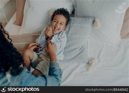 Sweet little boy kid laughing loudly and smiling while his loving mom tickling him on bed with lot of soft pillows, mother telling really funny story to son while spending happy time together at home. Sweet excited mixed race boy lying on bed and laughing while playing with his loving mom