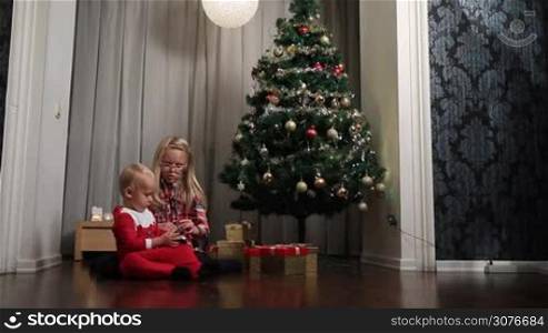 Sweet little girl and adorable toddler boy in Santa costume sitting on the floor under Christmas tree and opening presents. Cute boy playing with his toy car on the floor and smiling happy girl opening gift box.