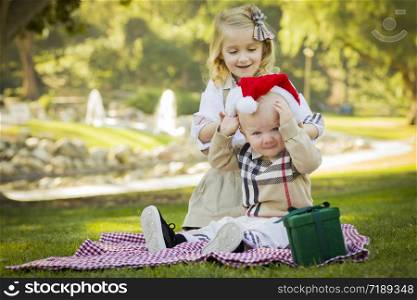 Sweet Little Girl Tries to Put A Santa Hat On Her Reluctant Baby Brother Outdoors at the Park.