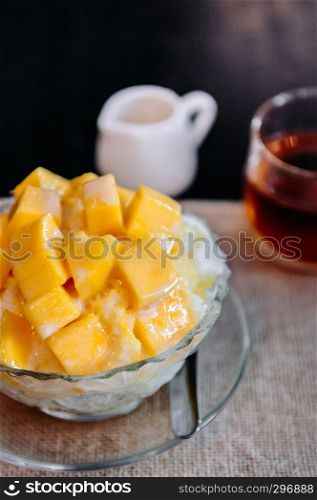 Sweet mango Binsu Korean shaved ice snow dessert in glass bowl with black tea, close up shot