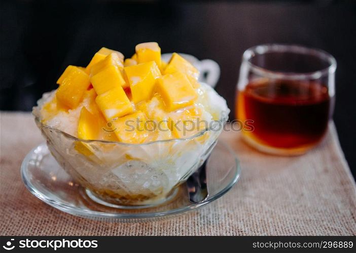Sweet mango Binsu Korean shaved ice snow dessert in glass bowl with black tea, close up shot