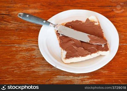 sweet sandwich from fresh toast with chocolate spread on white plate, table knife on wooden table