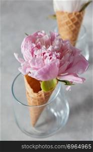 Sweet waffle cones with beautiful coloful peony flowers in glass cups on a gray background. Summer concept.. Delicate pink peony flower in a wafer cone in a glass standing on a gray stone table.