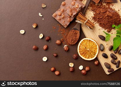 sweets, confectionery and culinary concept - chocolate with hazelnuts, cocoa beans, and powder with dry orange and cinnamon on wooden board with spoon on brown background. chocolate with hazelnuts, cocoa beans and powder