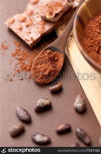 sweets, confectionery and culinary concept - chocolate with hazelnuts, cocoa beans and powder in wooden bowl with spoon on brown background. chocolate with hazelnuts, cocoa beans and powder