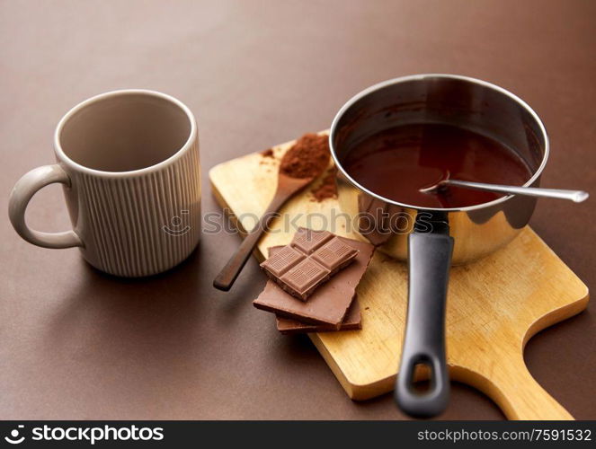 sweets, confectionery and culinary concept - pot with melted hot chocolate, cocoa powder in spoon, ceramic mug and wooden board on brown background. pot with hot chocolate, mug and cocoa powder